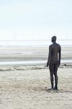 Another Place statue facing empty beach in Crosby, United Kingdom, with windmill power turbines in the far distance