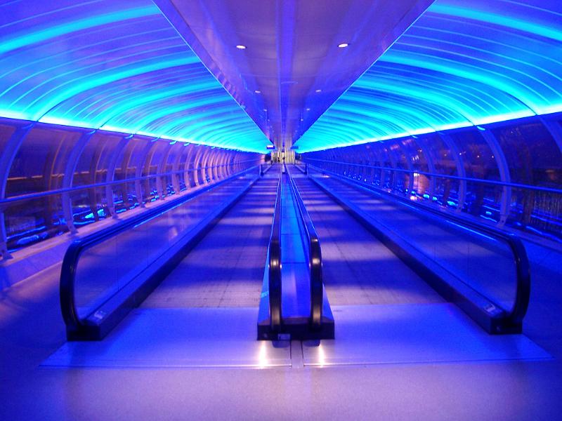 Interior of an airport walkway with two central moving travelators or passenger conveyor belts under a curved ceiling illuminated with blue lighting