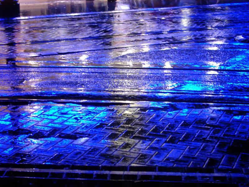 Rain falling on an urban sidewalk at night with blue reflections from lights in the layer of moisture for a colorful background