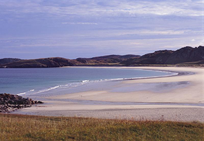 Sandy tranquil beach on the Isle of Lewis in the Hebrides with a curving shoreline and calm blue sea against a mountain backdrop