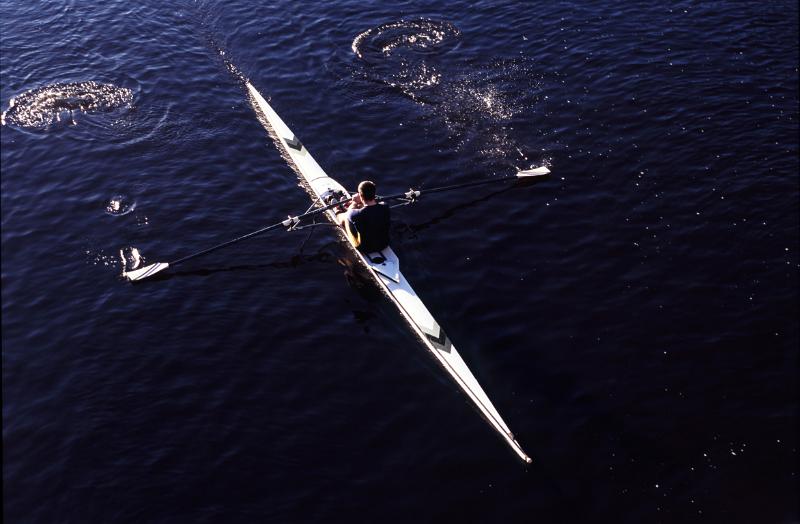 Overhead view of a man practising sculling in a single boat rowing along a river with water splash off the oars