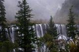 Scenic view of misty waterfalls in China cascading over a cliff amongst forested slopes and pine trees
