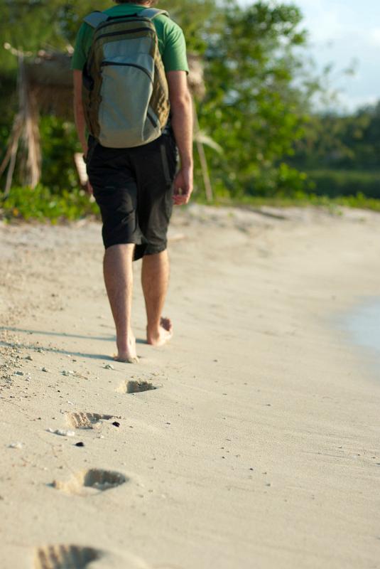 Man wearing a backpack walking away along a sandy beach leaving a trail of footprints