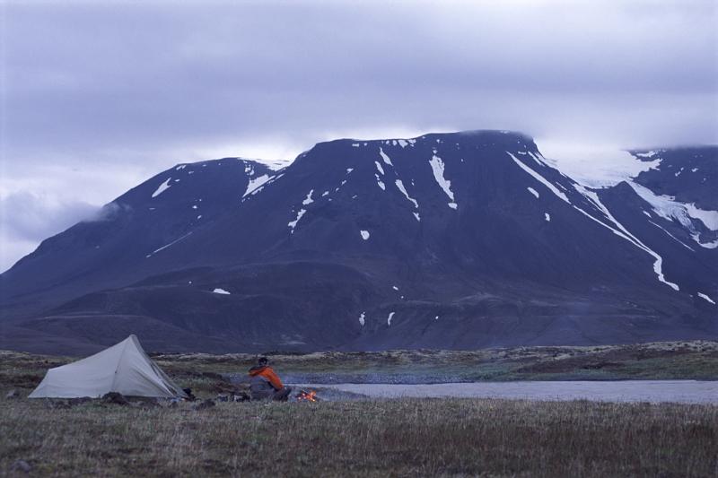 Camping in Iceland on the lowland plains below a snowy volcanic mountain range alongside a meltwater lake with a tent and person at a campfire