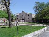 Grassy Landscape, Tall Trees and Tourists in Front Famous Historic Alamo Building. Isolated on Light Blue Sky Background.