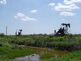Nodding Donkey, Nickname of Oil Well Drill Surrounded by Green Grasses and Flowing Water at Texas. Isolated on Light Blue Sky Background.