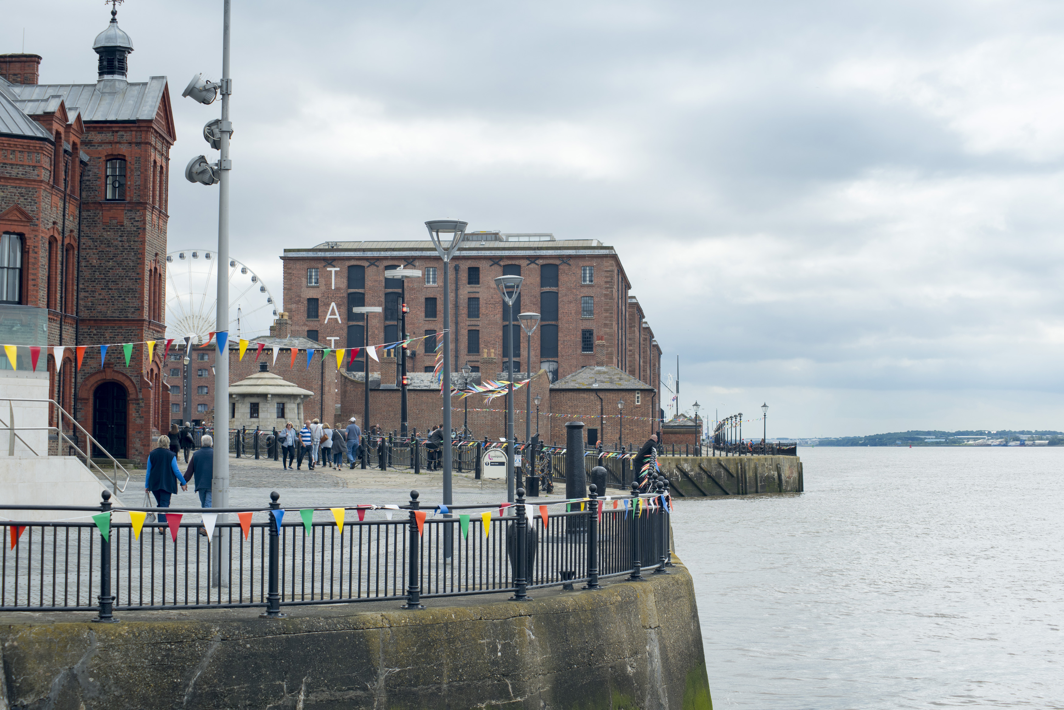 an image of Various tourists walking around landmark buildings near round railing along waterfront at Liverpool Mersey