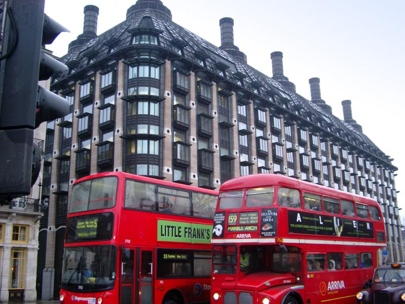 Red London Buses in Tour with Architectural Big European Building on the Street Side.