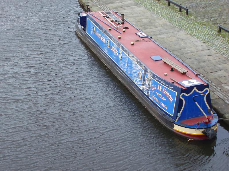 High angle view of a traditional English canal narrow boat with advertising signs on the side moored at the side of the canal alongside a paved walkway