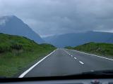 View through the windshield of a car of a tarred road through the Scottish highlands with high mountain peaks on a cold overcast day
