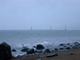 View from a rocky beach of three yachts sailing offshore on the horizon on a cold bleak misty day