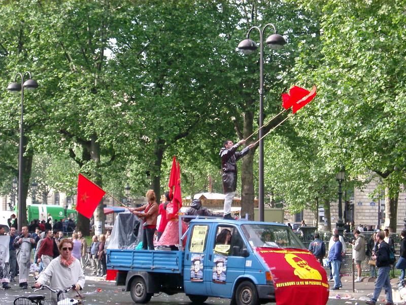Men with Flags on Vehicle with Random People on Ground During May Day Parade. Isolated on Tall Green Trees.