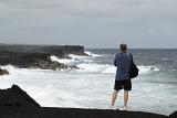 Tourist standing with his back to the camera on the black volcanic sand on Kalapana beach, Hawaii looking out over the ocean waves and rocks